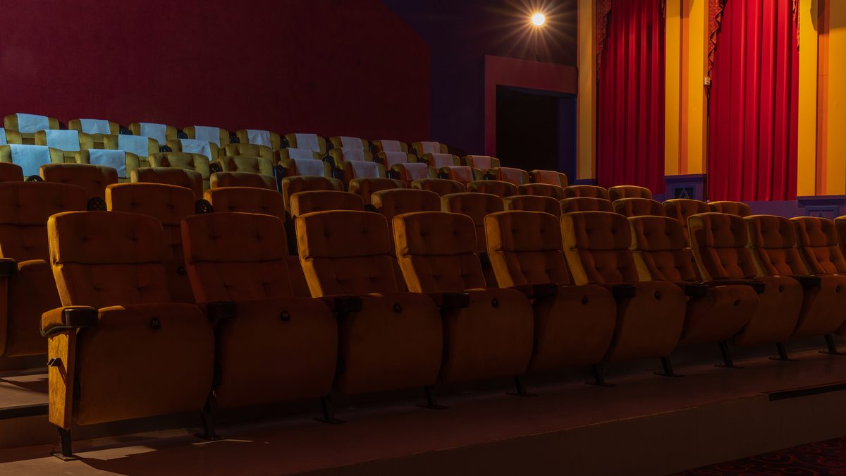 Rows of empty mustard-coloured seats in a cinema hall, with red curtains beside the screen wall
