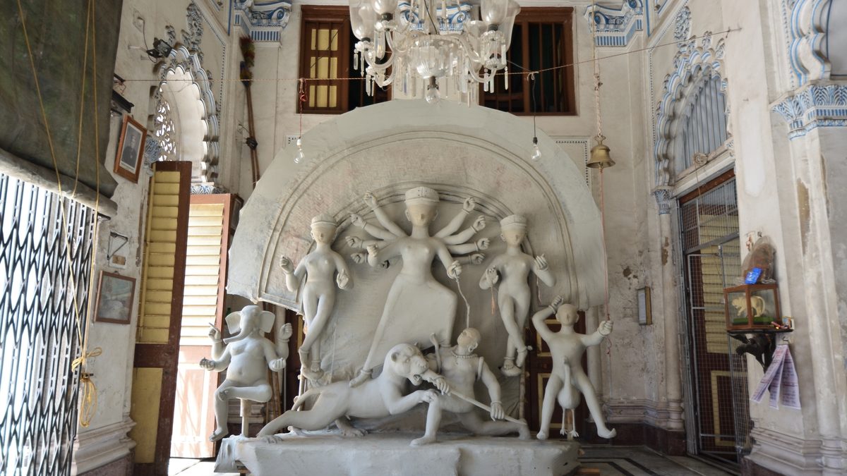 An unpainted clay Durga with Lakshmi, Saraswati, Ganesh, Kartik, the lion and Mahishasura under one arched frame, in a thakurdalan with blue plaster work and a glass chandelier