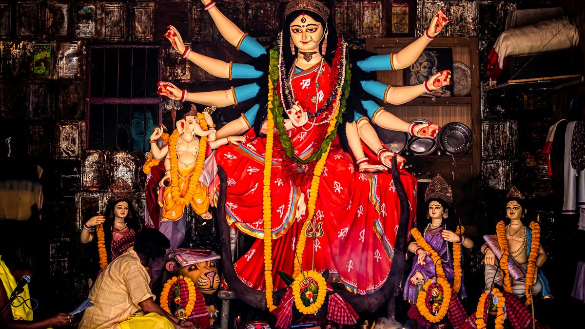 A Durga Puja mandap: a hand holding the mantra text to a microphone at the left edge, a man in a kurta setting out offerings, before a Durga in a red sari with Ganesha, Lakshmi, Saraswati and Kartik