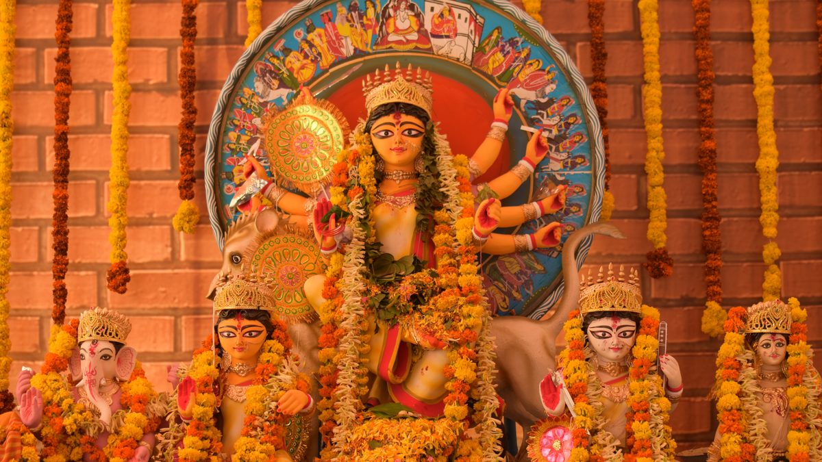 Durga with Lakshmi, Saraswati, Ganesh and Kartik under a painted chalchitra, heaped with marigold and rajanigandha garlands, against a brick wall at a Bengaluru puja