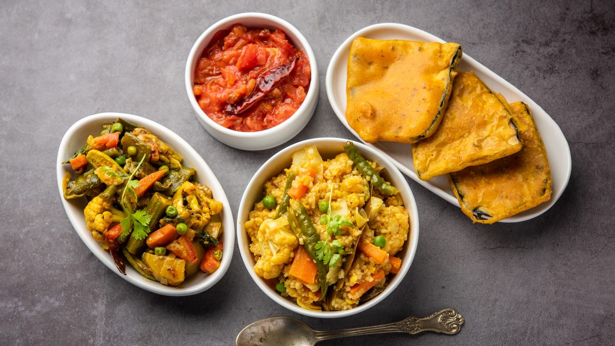Three white bowls and a long white plate on a grey table, seen from above: khichuri with cauliflower, peas, carrot and beans, labra, a sweet tomato chutney with a dry red chilli, and slices of beguni, with an old tarnished spoon