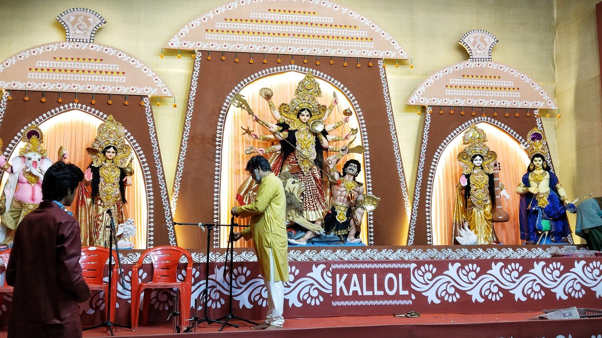 A Durga Puja stage in Mumbai: Durga with her children under three painted arches, a man in a yellow kurta setting up microphones in front, red plastic chairs and a banner reading Kallol