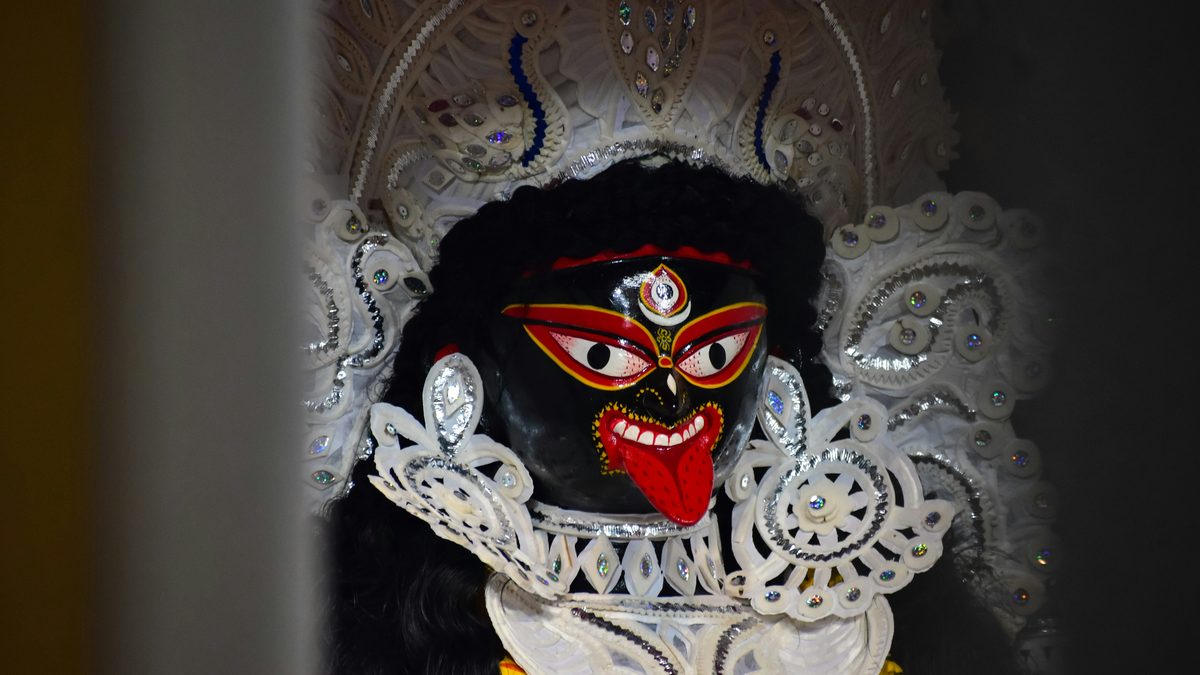 পশ্চিমবঙ্গের এক কালো কালীপ্রতিমার মুখ: লাল জিভ বার করা, লাল-হলুদে আঁকা তিন চোখ, মাথায় সাদা শোলার মুকুট