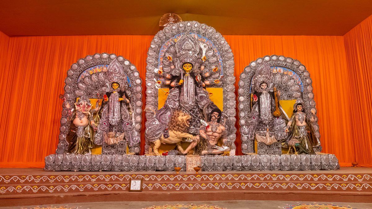An orange-draped mandap with the whole Durga family under three silver arches: Ganesha and Lakshmi on the left, Durga on the lion with Mahishasura in the middle, Saraswati and Kartik on the right, alpana on the floor in front