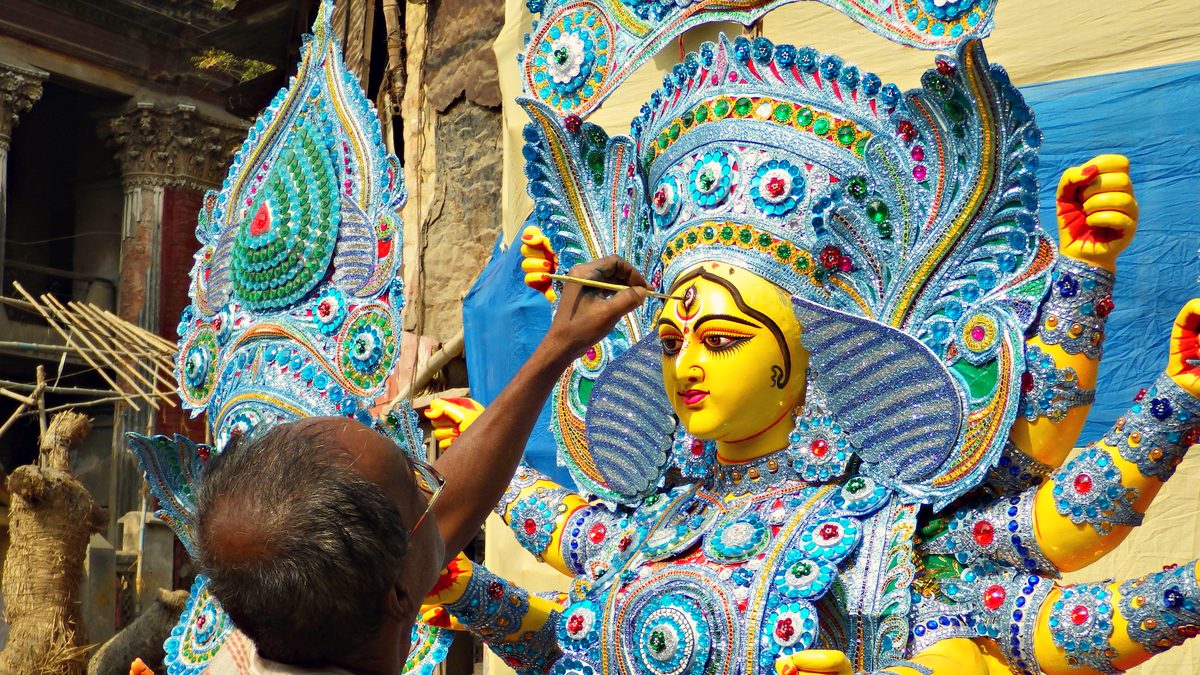 An elderly craftsman, seen from behind, paints the eyebrow of a yellow-faced goddess covered in blue and silver sequin work, in an open workshop with bamboo poles and an old pillared house behind