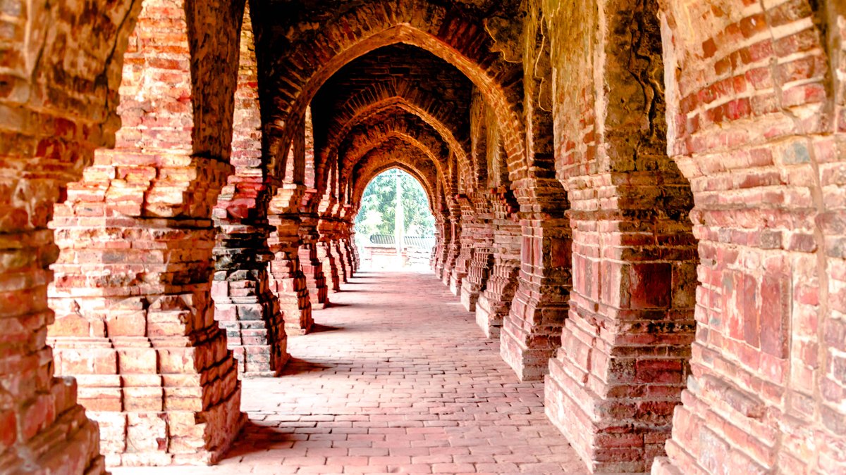 A long corridor of red-brick arches at a terracotta temple in Bishnupur, each arch framed inside the next, with green trees in daylight at the far end
