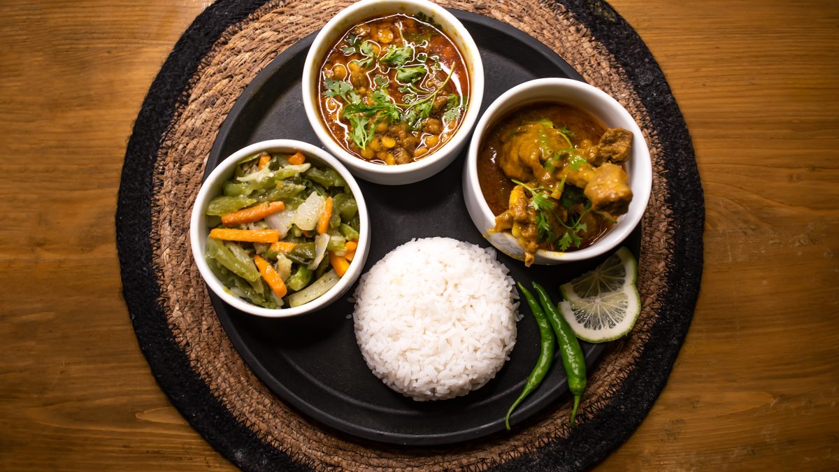 A black round plate on a jute mat, seen from above: a mound of white rice, bowls of chicken rezala, chana dal cooked with meat and mixed vegetables, two green chillies and a slice of lime