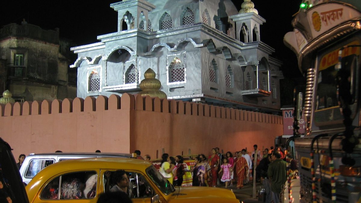 A lit pandal shaped like a domed palace behind a pink crenellated wall in Ahiritola, with a yellow Ambassador taxi and a crowd of families walking past in the small hours
