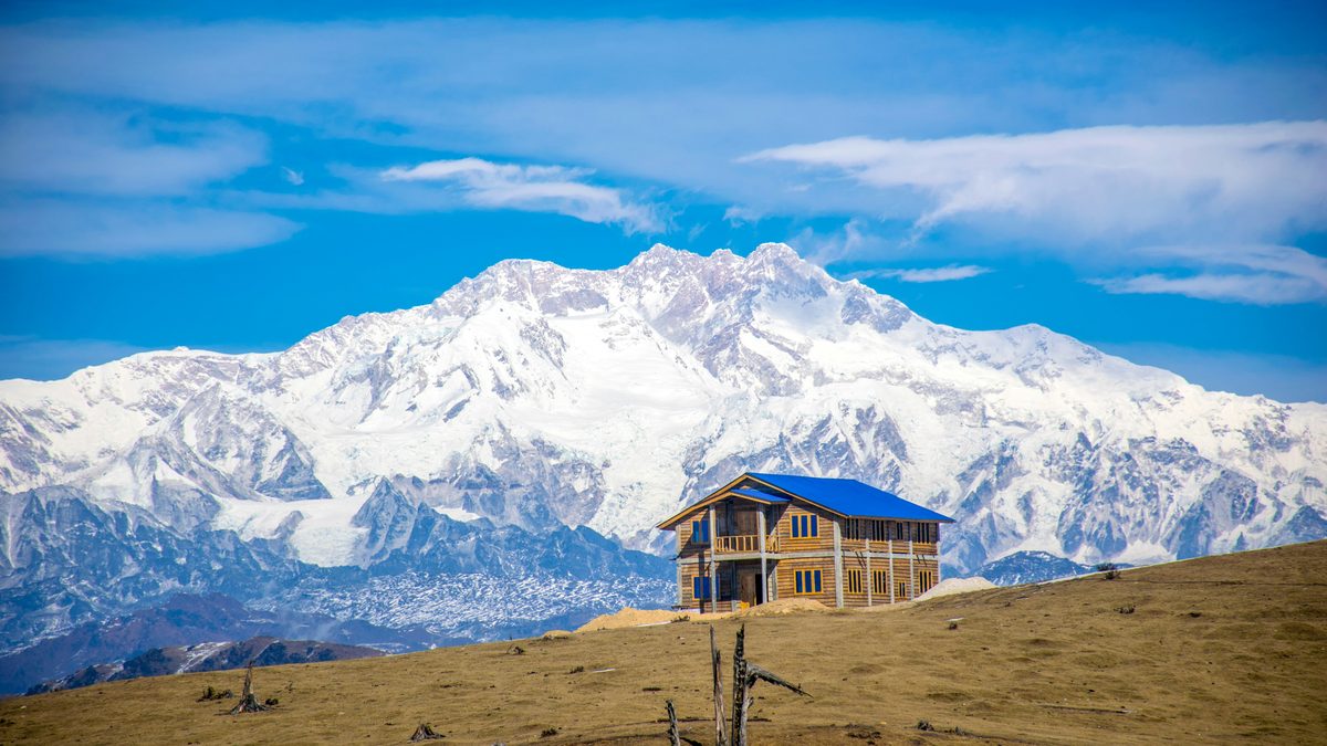 A wide range of snow-covered peaks under a blue sky with thin white cloud. In front, a brown grassy slope with a two-storey timber building under a blue roof, and a bare dead tree and a few stumps in the foreground
