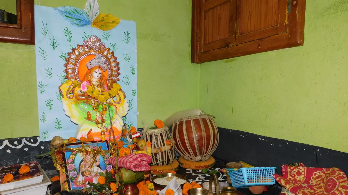 বাড়ির সরস্বতী পুজো: সবুজ দেওয়ালের সামনে রং করা সরস্বতী প্রতিমা, গাঁদাফুলের নীচে বইয়ের গোছা, এক জোড়া তবলা আর পেতলের বাসন