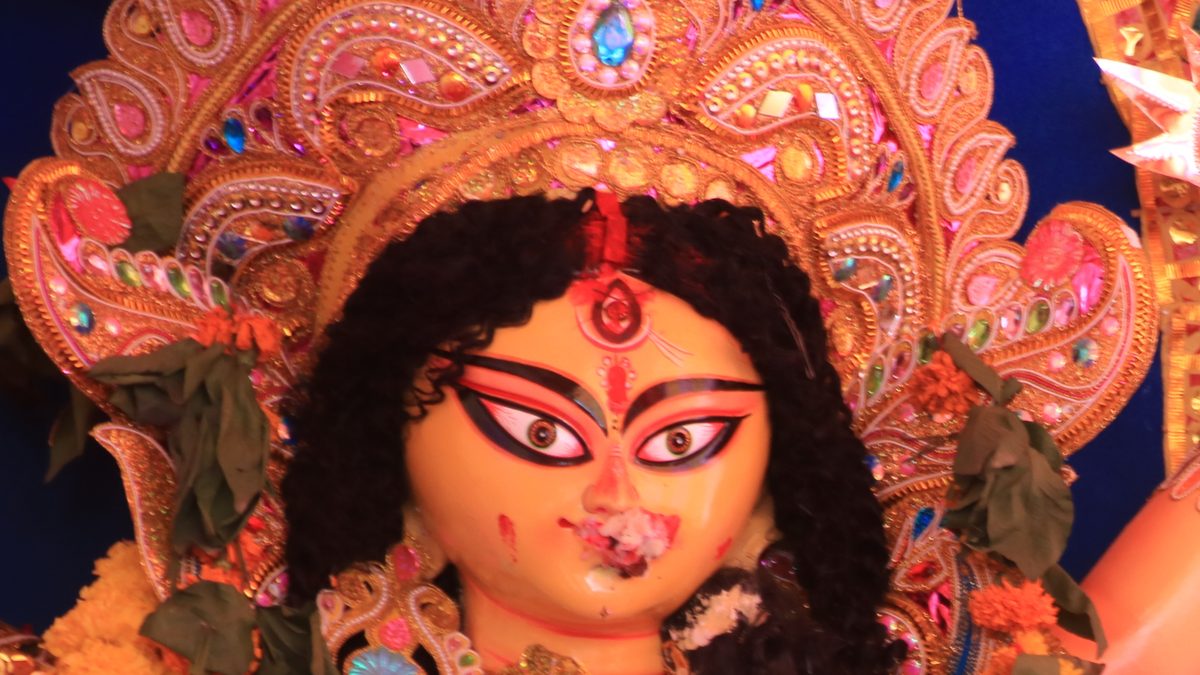 Close-up of a Durga idol on Bijoya Dashami, sindoor at her parting and forehead and a sweet pressed to her lips, framed by a gold and pink crown and halo